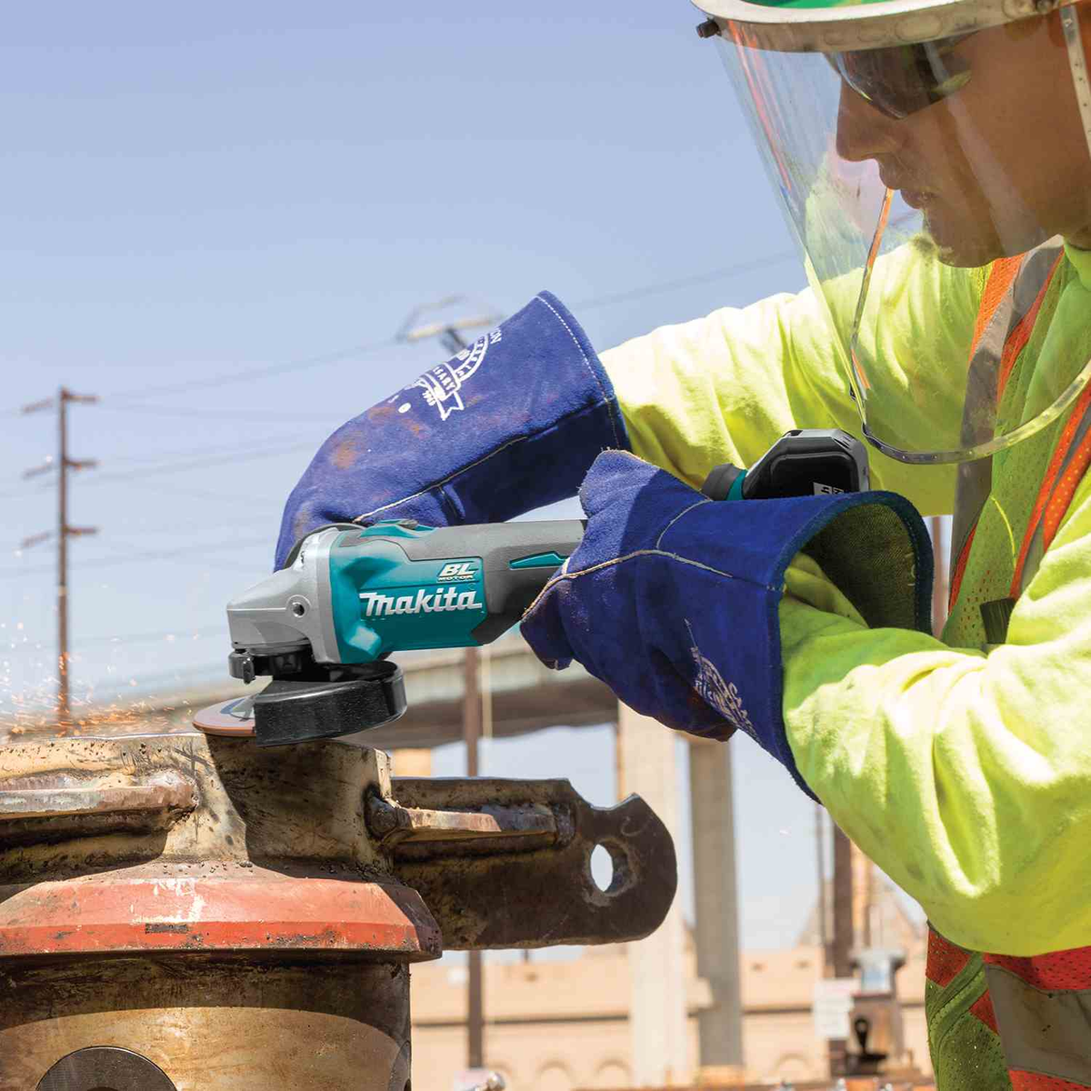 A construction worker wearing a clear face shield and high-visibility safety jacket uses a teal and gray Makita angle grinder on a metal pipe at an outdoor construction site. Get yours today at B&R Lawnmower Shop, 20032 Saticoy Street, Winnetka, CA 91306. Near Canoga Park on the intersection of Winnetka and Saticoy. Serving Los Angeles County and San Fernando Valley since 2001.