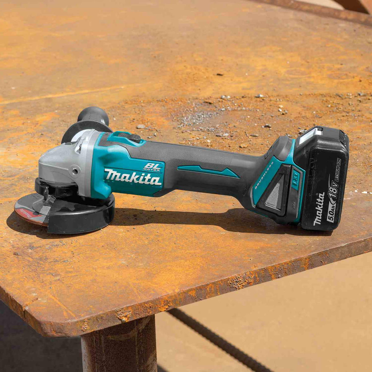 A Makita cordless angle grinder with a black and teal 5.0 amp hour Makita LXT 18V battery pack, resting on a rusty metal workbench. Get yours today at B&R Lawnmower Shop, 20032 Saticoy Street, Winnetka, CA 91306. Serving Los Angeles County and San Fernando Valley since 2001.