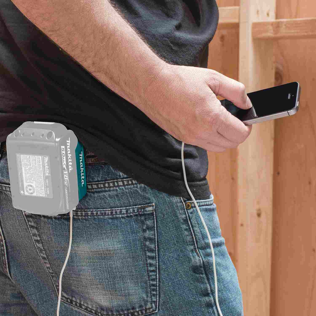 Person charging a smartphone with a Makita LXT 18V battery pack attached to an ADP05 power source, standing in a construction job site.