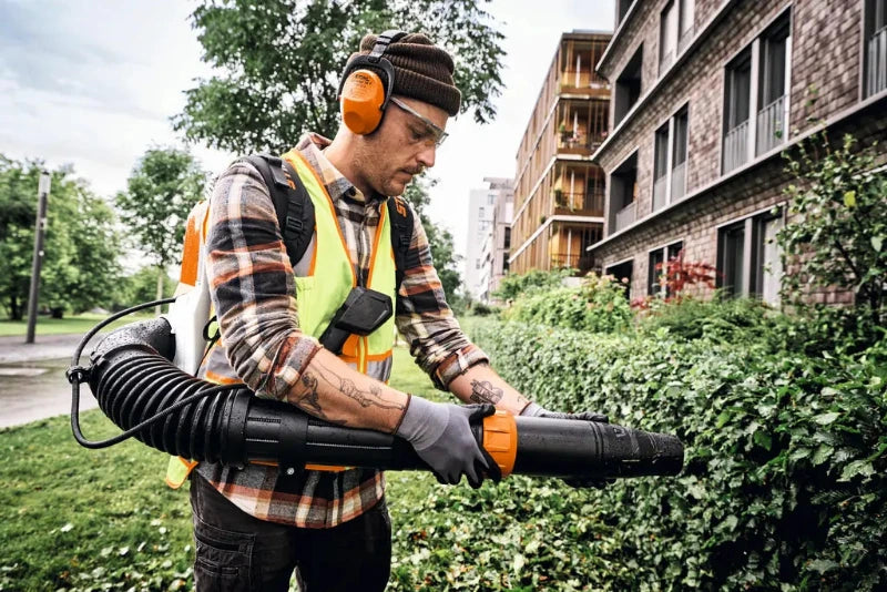 A man wearing safety gear, including orange earmuffs, a high-visibility vest, and gloves, operates a black and orange STIHL BRA 280 battery-powered STIHL AP/ALLPRO leaf blower outdoors in front of a brick apartment building and green bushes.