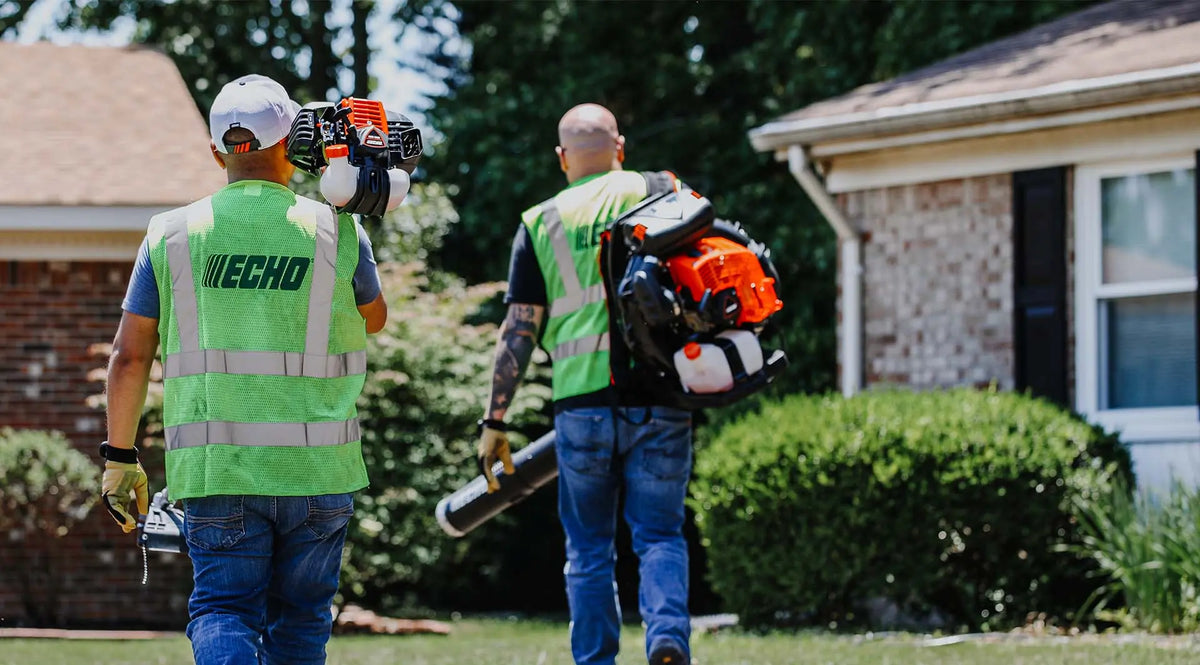 Two men wearing high-visibility Echo safety vests walk outdoors in a yard while carrying Echo leaf blowers on their backs.